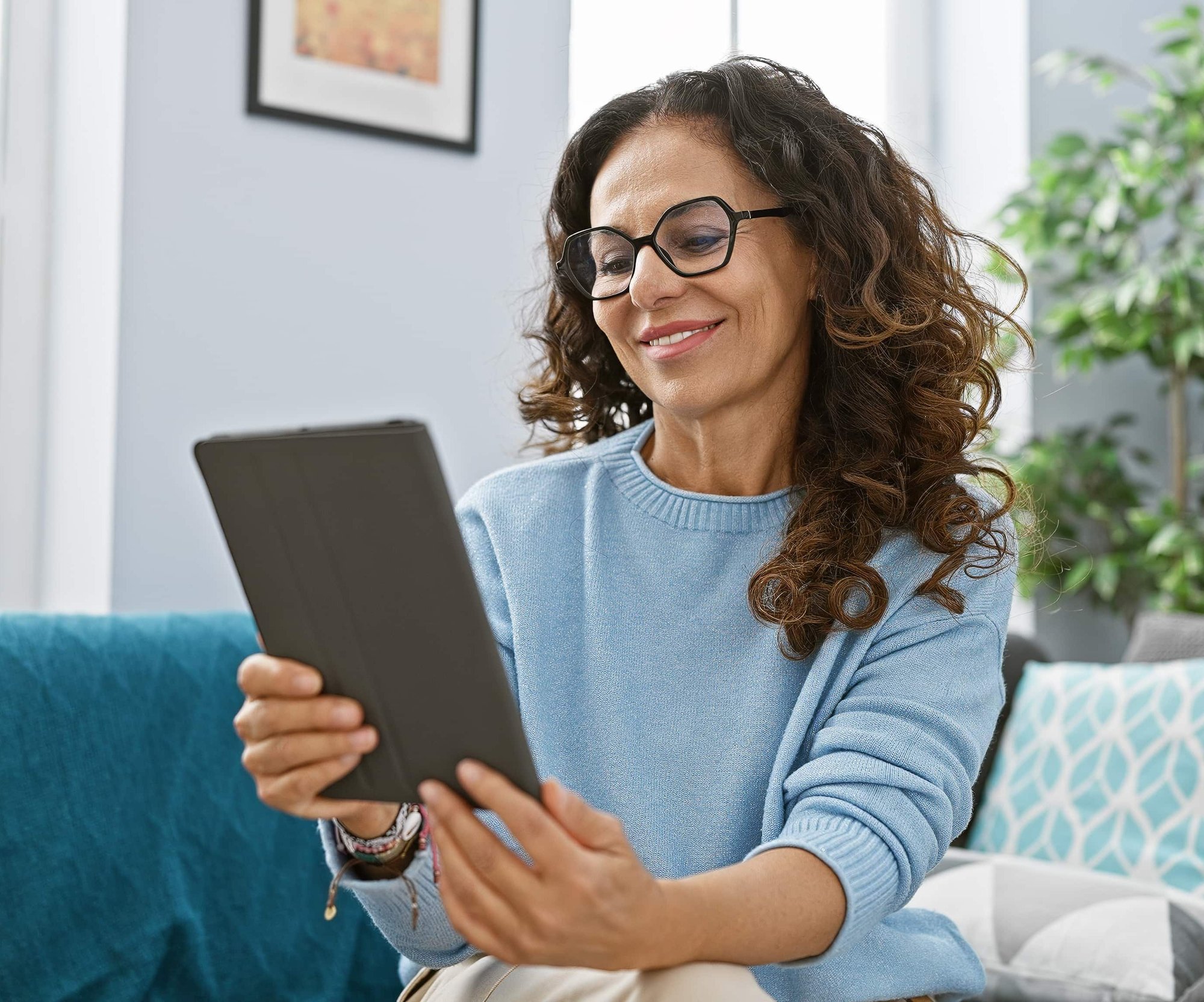 Smiling woman with curly hair wearing glasses and blue sweater using a tabletSmiling woman with curly hair wearing glasses and blue sweater using a tabletSmiling woman with curly hair wearing glasses and blue sweater using a tablet_873455312-min-1